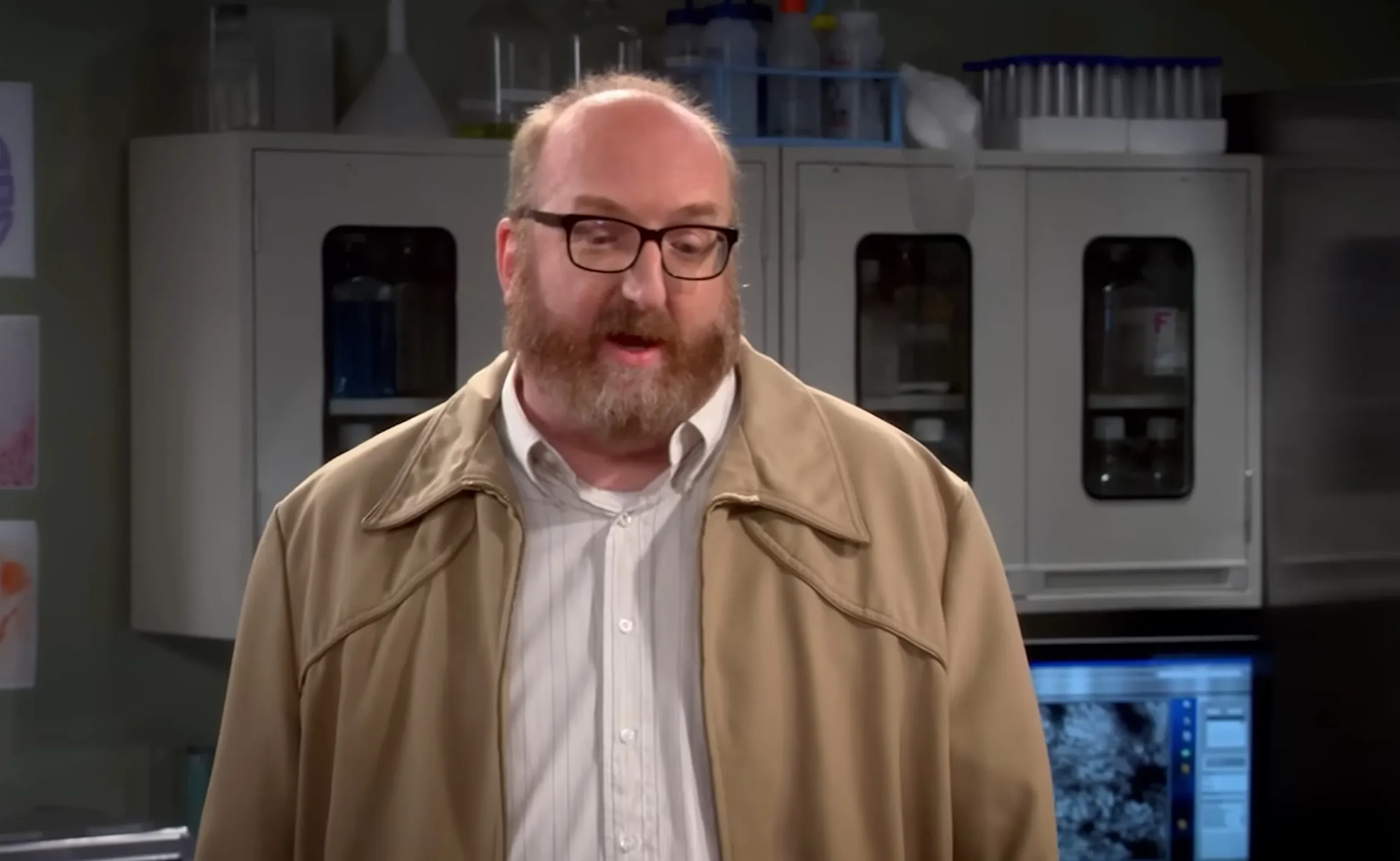 Bert Kibbler from The Big Bang Theory wearing glasses and a beige jacket standing in a lab setting.