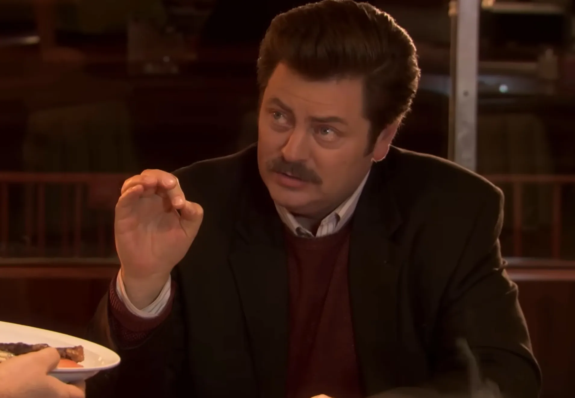 Side character from Parks and Recreation sitting at table and gesturing with his hand during conversation indoors.