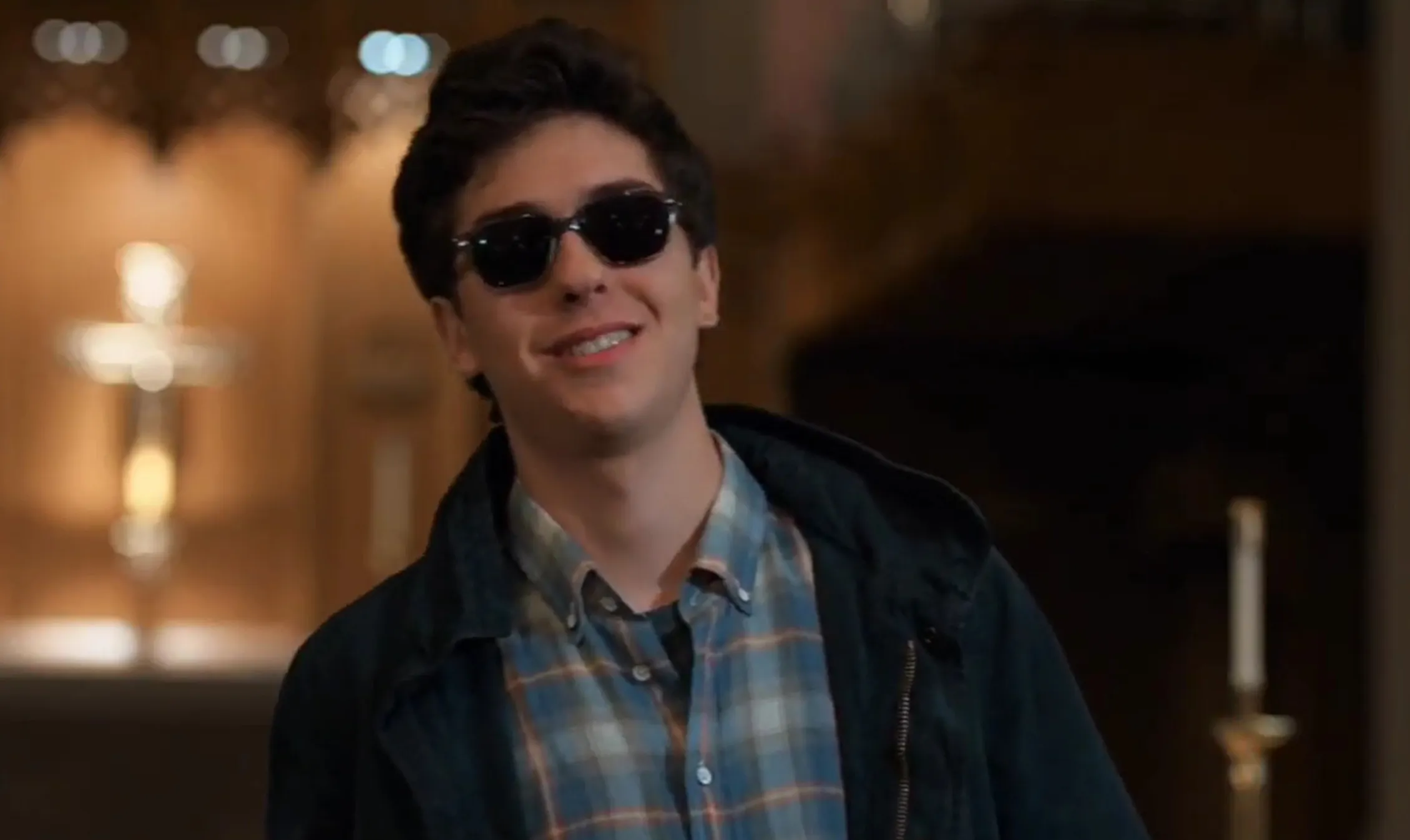 Supporting character from The Fault in Our Stars wearing sunglasses and a plaid shirt indoors with blurred background lighting.