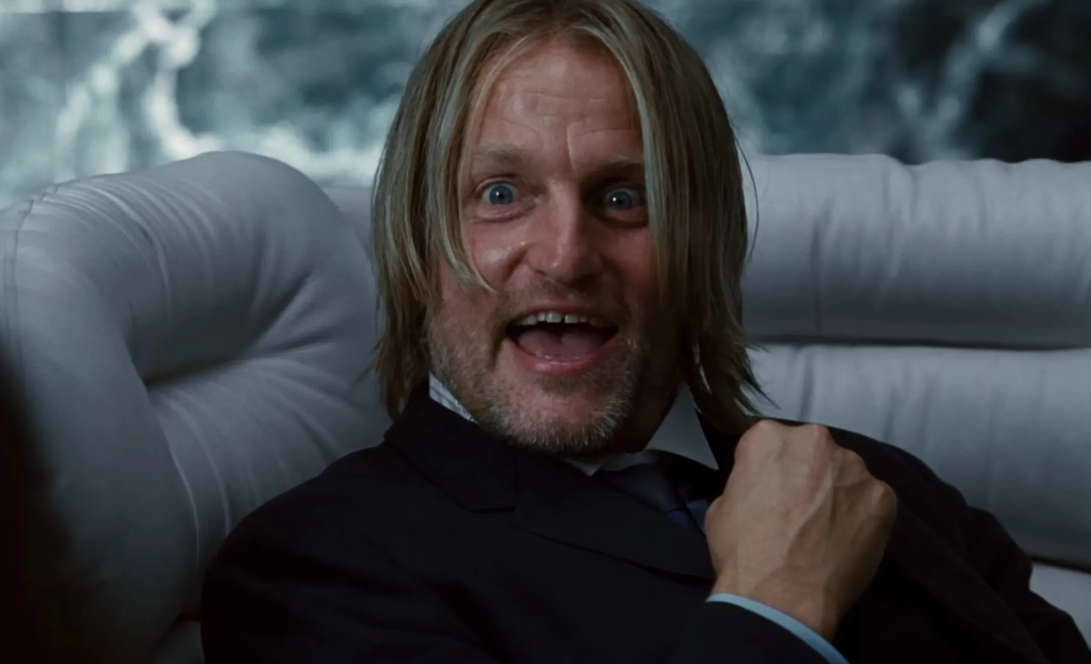 Supporting character from The Hunger Games with long hair and a beard, sitting on a white couch, animated and expressive.