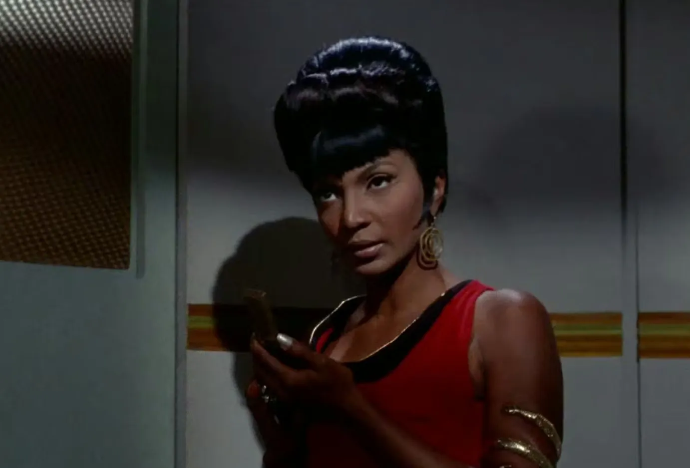 Woman in a red dress from Star Trek The Original Series holding a device with styled black hair and gold accessories.