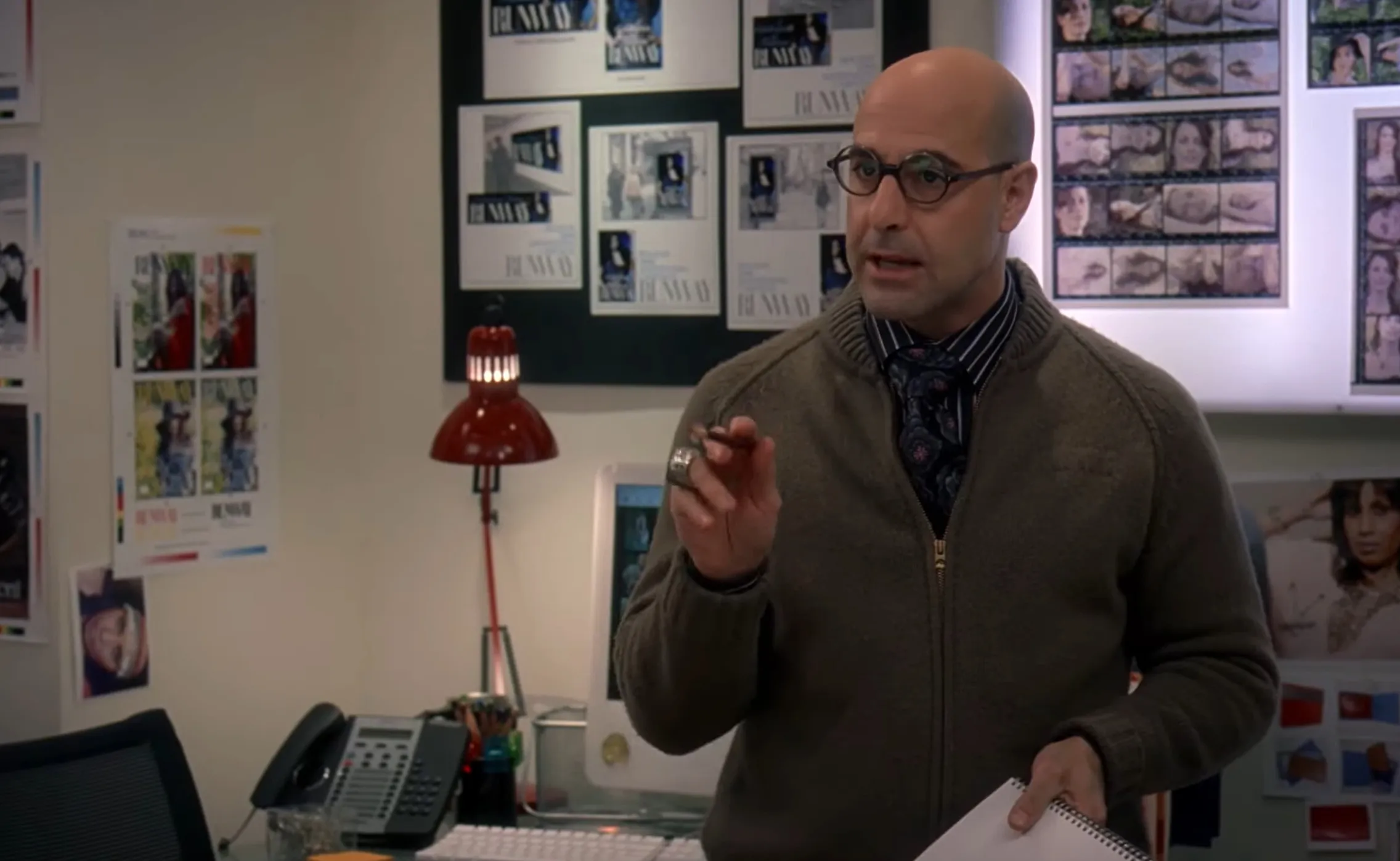 Supporting character wearing glasses and a brown sweater speaking inside an office with posters on the walls