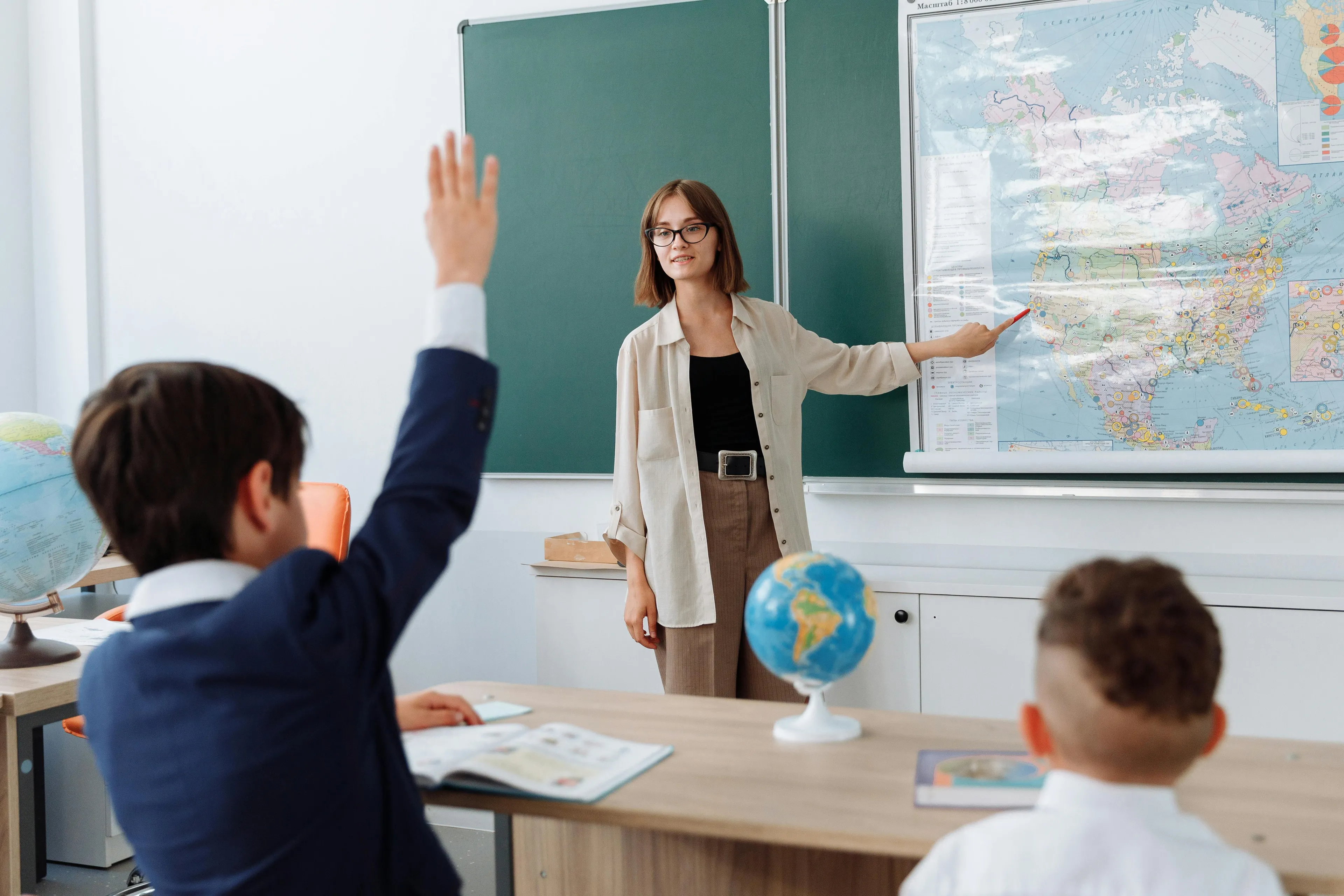 Teacher pointing at a map during a geography quiz, with students engaged and one raising a hand to answer.