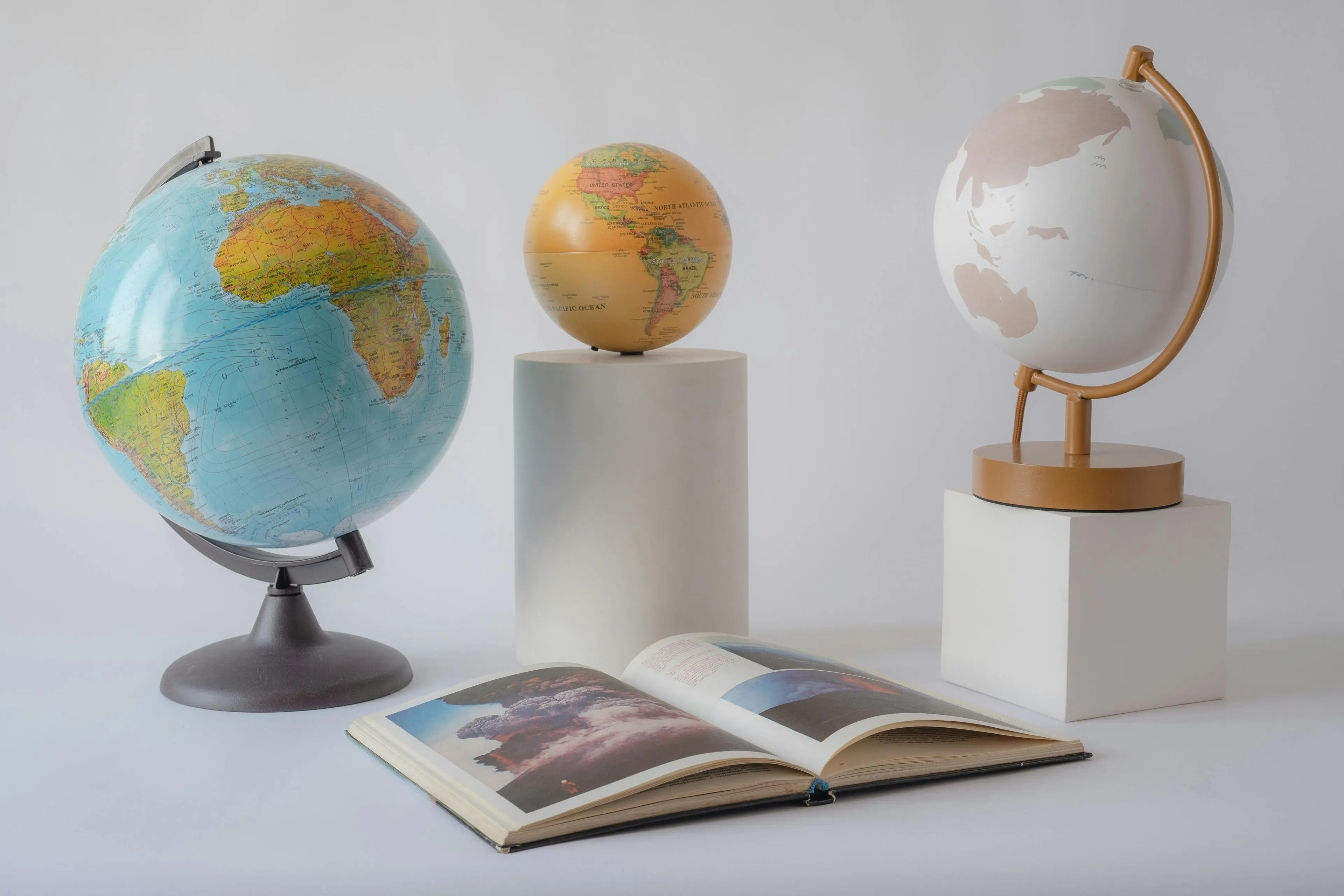 Three different globes and an open geography book displayed on white pedestals in a minimalistic setting.