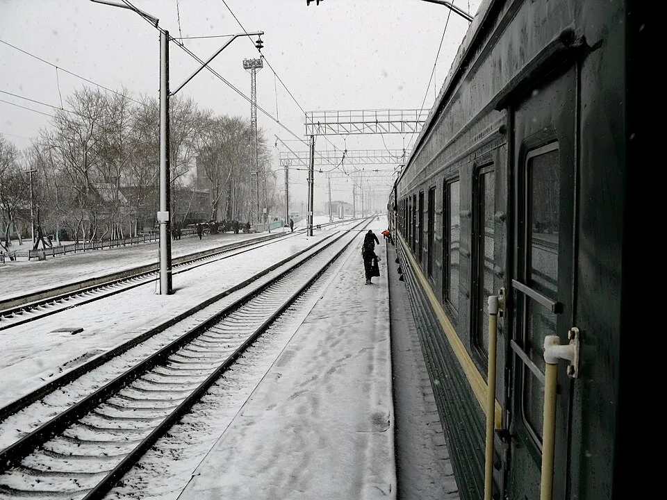 View from a train on the Trans-Siberian Railway at a snowy station with tracks and passengers visible.