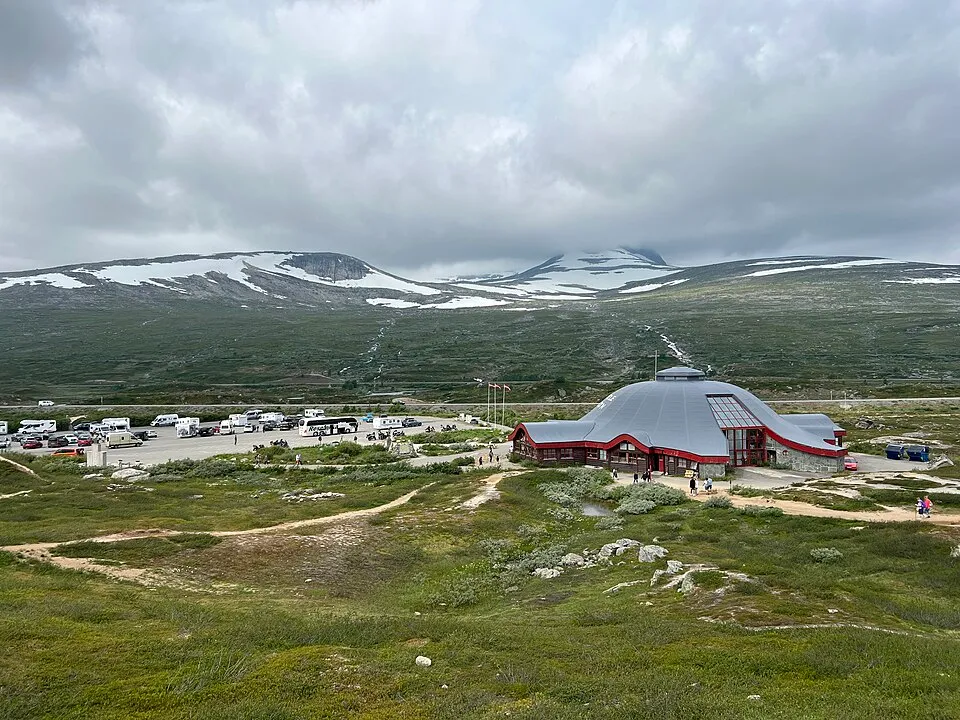Visitor center and parking lot in northern Norway above the Arctic Circle with snowy mountains under summer clouds.