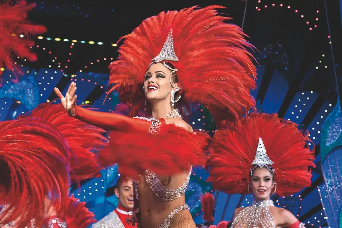 Showgirl dancers in vibrant red costumes performing at a tourist attraction with bright, colorful stage lighting and decorations.