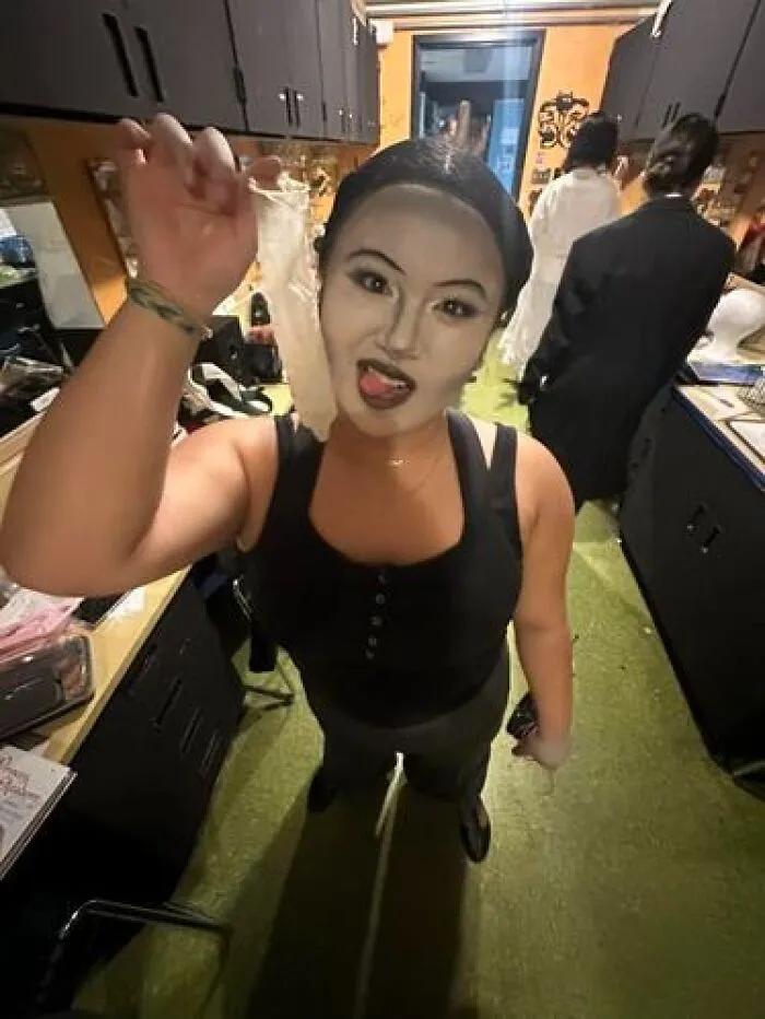 Person with magical stage makeup in a backstage dressing room holding a peeled face prosthetic piece and sticking out tongue.