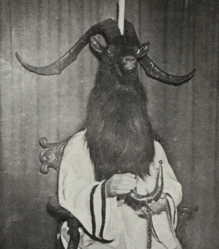 Old black and white creepy photo of a person wearing a goat head mask with large curved horns seated on a chair.