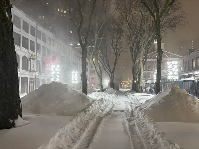 Snowstorm hitting a U.S. city street at night with heavy snow accumulation on sidewalks and trees lining the path.