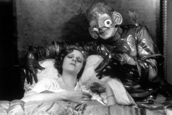 Old black and white creepy photo of a person in a strange costume looming over a sleeping woman, odd and eerie scene.