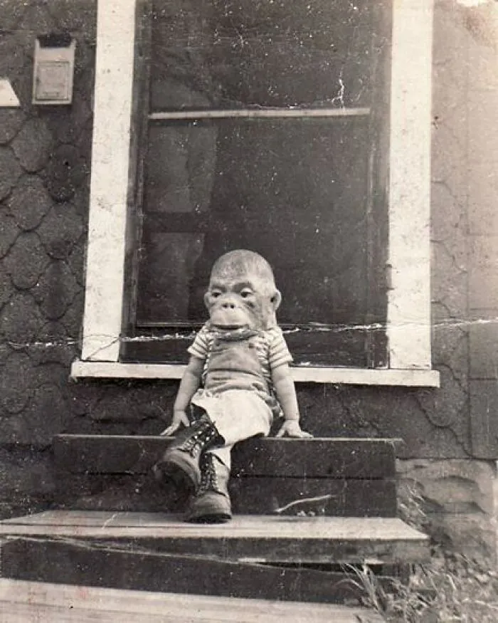 Vintage black and white photo of a child wearing a creepy ape mask sitting on wooden steps outdoors.