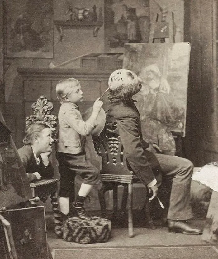 Old black and white creepy photo of a child painting an eerie face on a seated man's head in a vintage room.