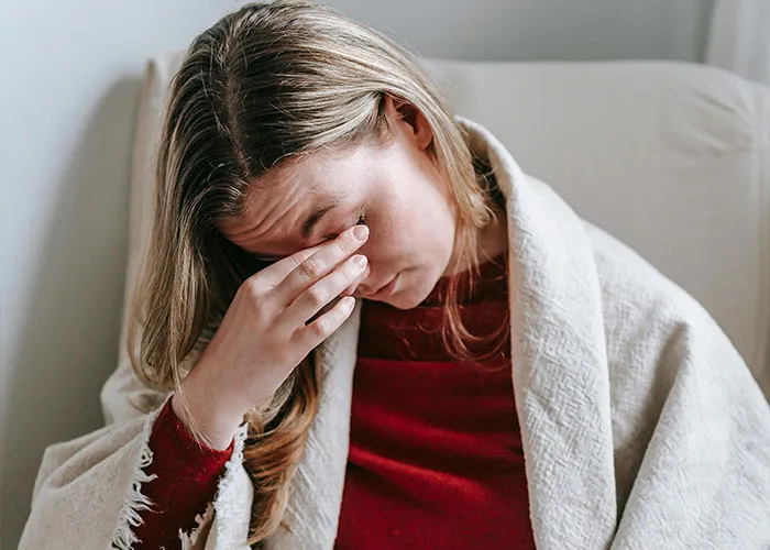 Woman wrapped in blanket holding her head, appearing unwell, illustrating medical issues that are difficult to endure.