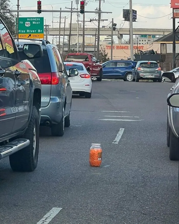 A jar filled with orange cheese balls found abandoned in the middle of a busy street among parked cars.