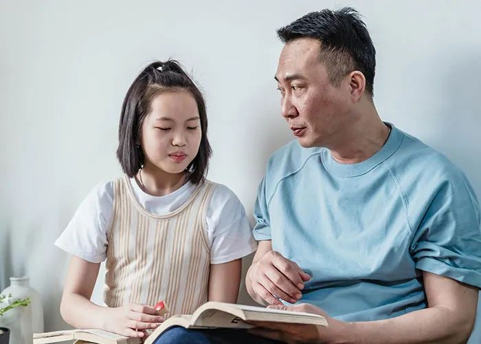 Asian man and young girl reading a book together, depicting emotions of discovering he is not the biological parent.