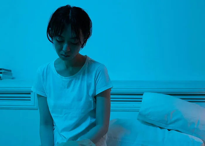 Woman in white shirt sitting on bed, looking distressed, illustrating medical issues nobody wishes to face.
