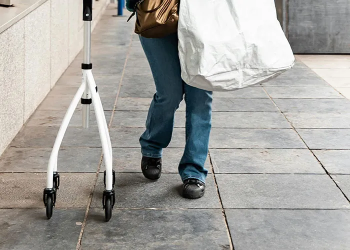 Person walking with a white walker and carrying a large bag, illustrating struggles from medical issues.