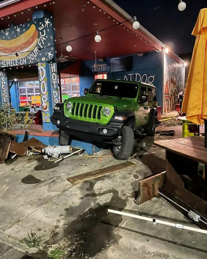 Green Jeep crashed into the corner of a colorful hot dog restaurant with debris scattered on the street at night.