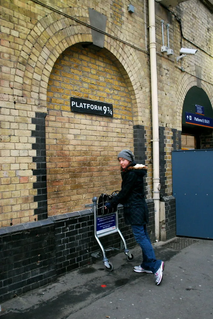 Person posing at the famous Platform 9 3/4 tourist attraction with luggage trolley halfway into the brick wall.