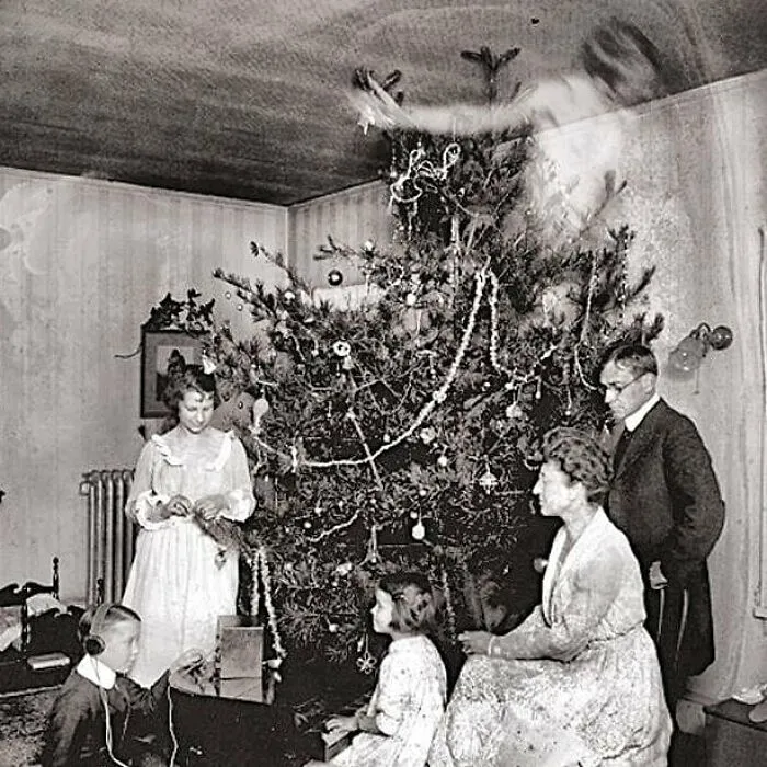 Old black and white photo of a family by a Christmas tree with a ghostly figure floating above, evoking odd and creepy vibes.