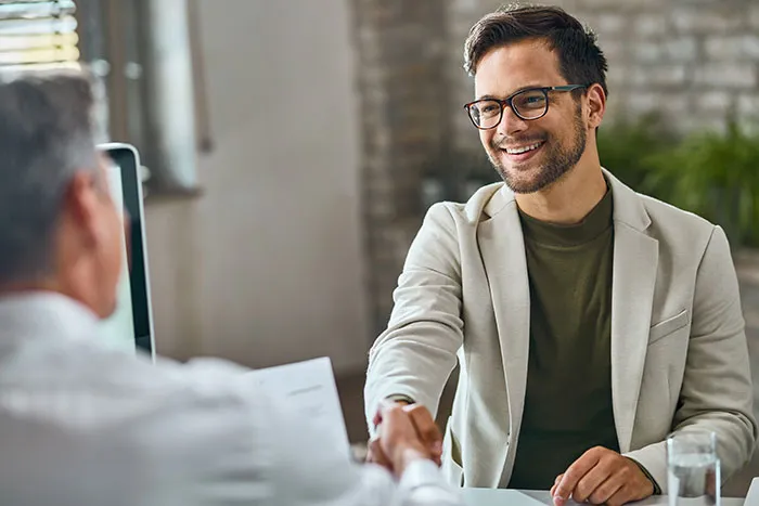 Young job candidate smiling and shaking hands with recruiter during interview in a modern office setting.