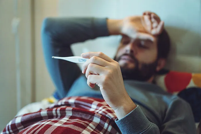 Man lying in bed covered with a blanket, checking his temperature, illustrating things about men guys want women to understand.