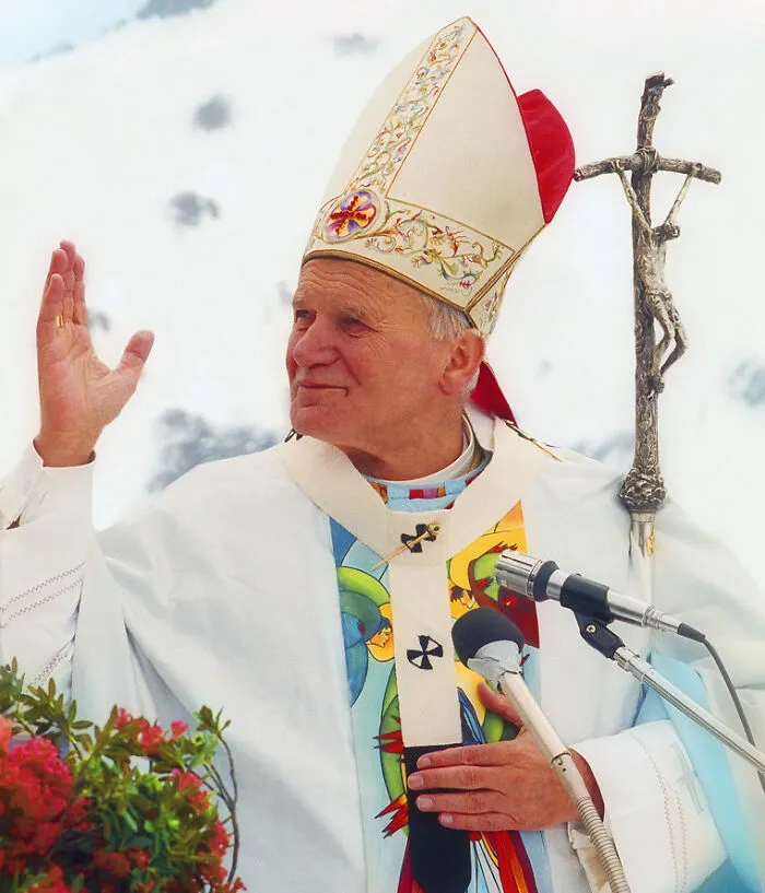 Religious leader in ceremonial attire speaking at a microphone with local inside jokes shared by people in the background.