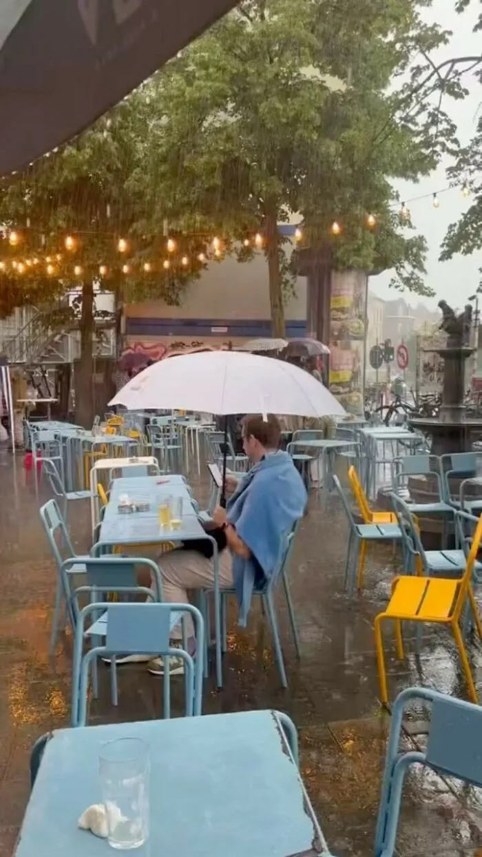 Man sitting under umbrella at outdoor café in the rain, showcasing weird and unhinged things on Brussels streets.