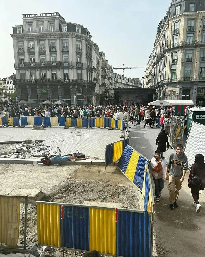 Person lying on a construction site surrounded by barriers amid a busy crowd on the streets of Brussels.