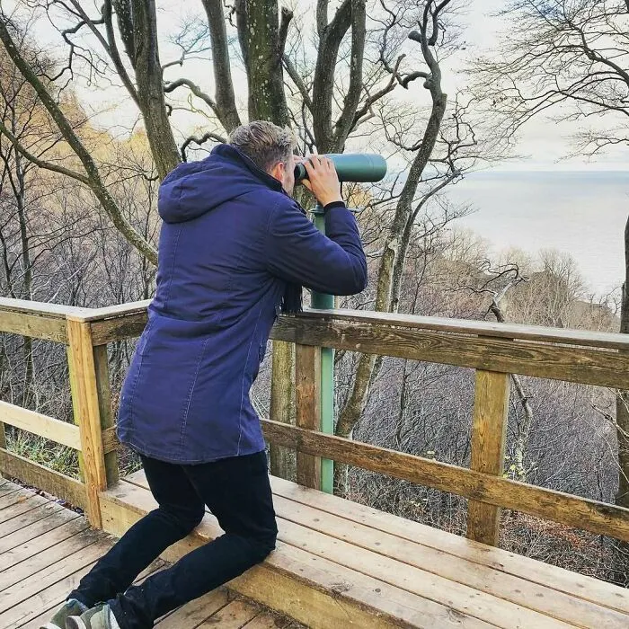 A tall person in a blue jacket leaning over a wooden railing to look through binoculars at a wooded outdoor viewpoint.