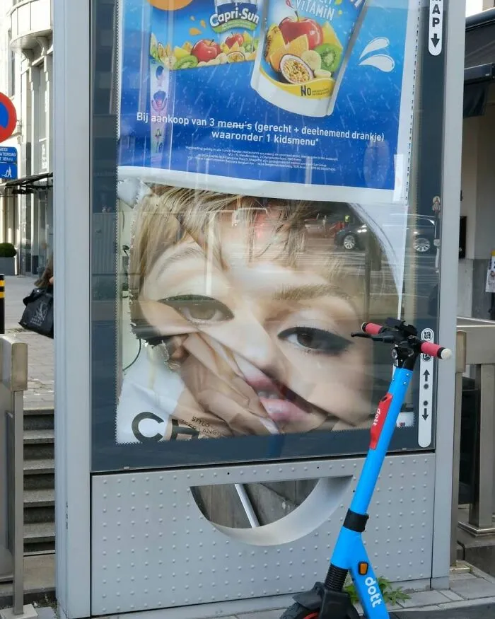 Distorted street advertisement of a face behind glass with reflections, electric scooter parked nearby on Brussels street.