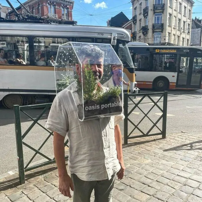 Man wearing a clear portable oasis planter on his head among buses on the streets of Brussels.