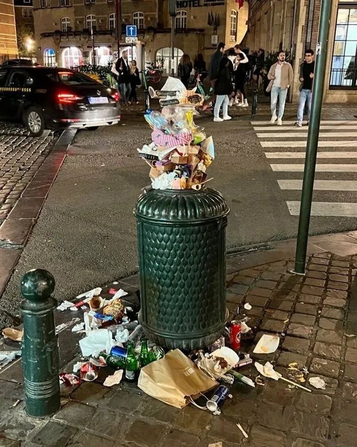 Overflowing trash bin with litter scattered around on a busy street in Brussels, showcasing weird and unhinged street scenes.