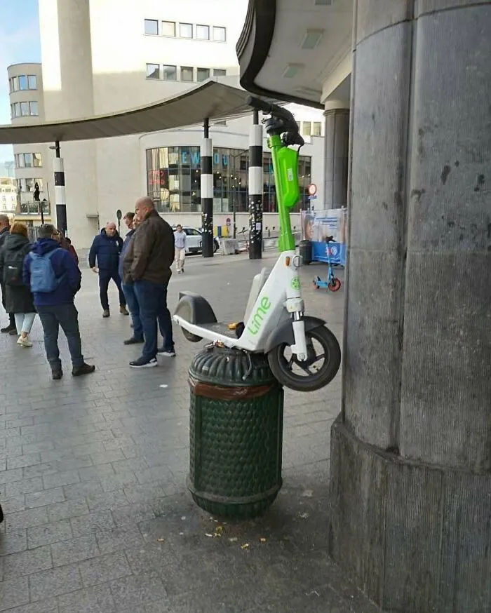 Lime scooter unhinged and bizarrely balanced on top of a trash can on the streets of Brussels with people walking nearby.