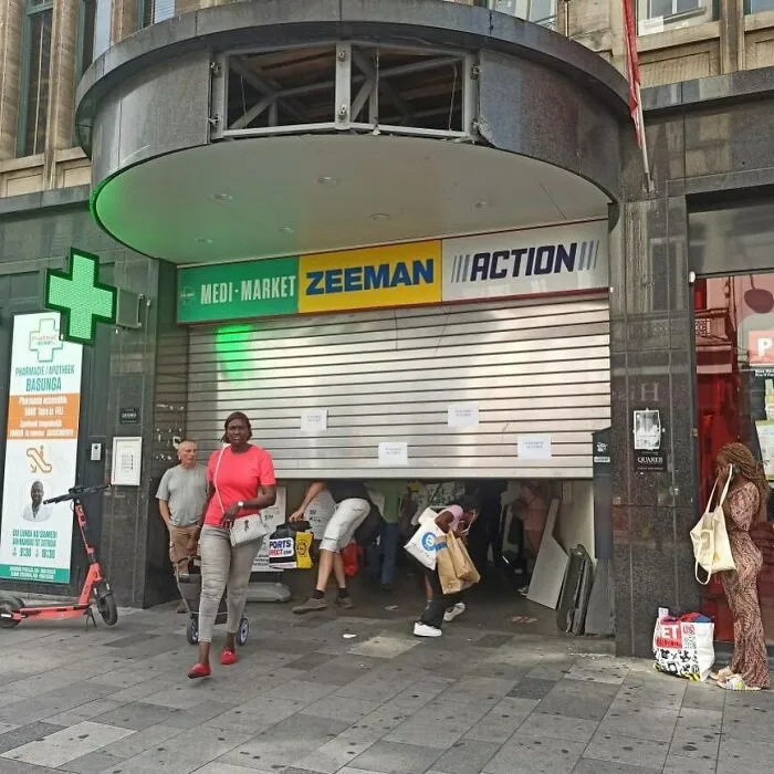 People squeezing under a partially closed storefront shutter on the streets of Brussels, showcasing weird street scenes.