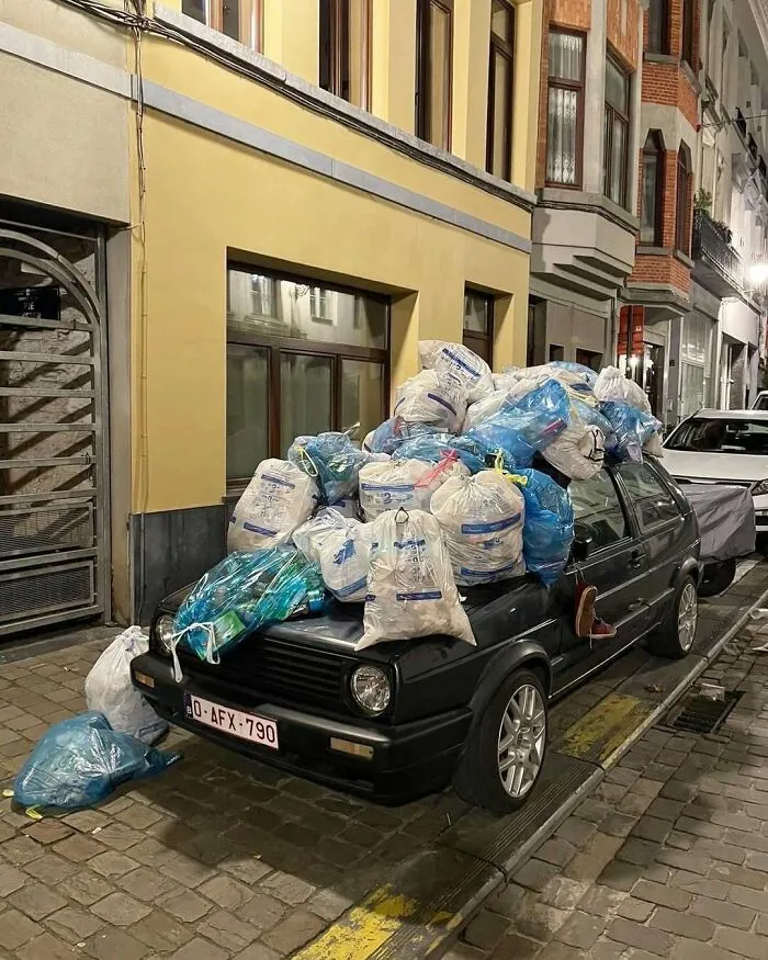 Black car on a narrow Brussels street overloaded with numerous white and blue garbage bags stacked on its roof and hood.