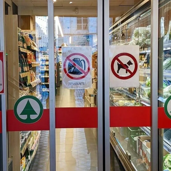 Store entrance in Brussels with unusual signs prohibiting dogs and students carrying backpacks inside.