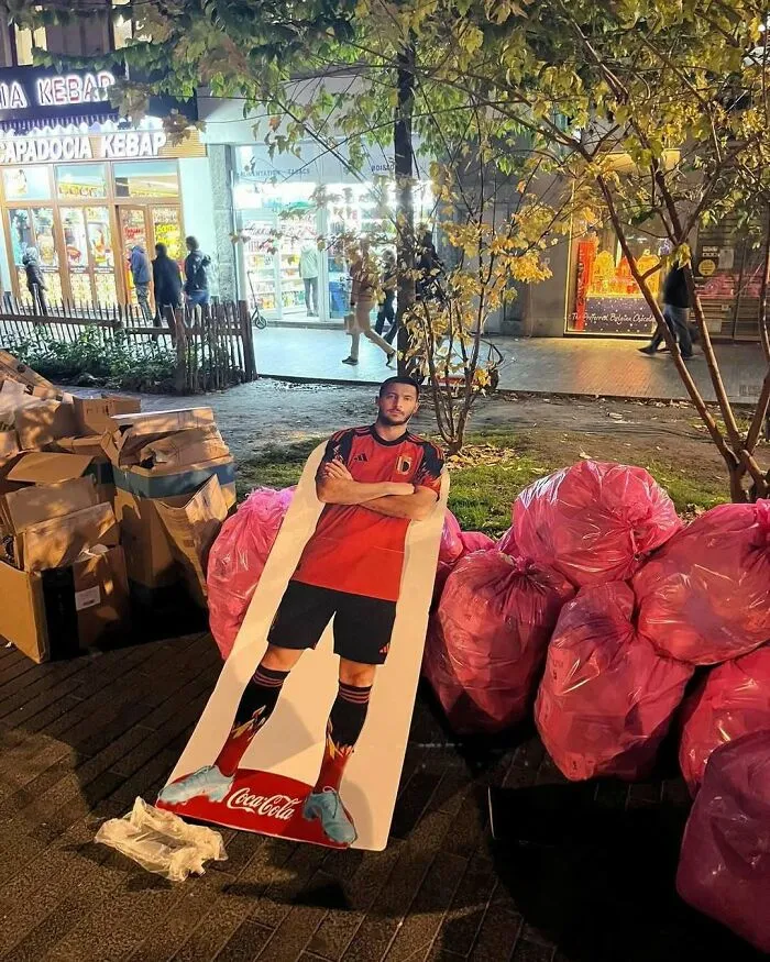 Cutout of a soccer player in a Coca-Cola ad discarded beside pink trash bags on the streets of Brussels at night.