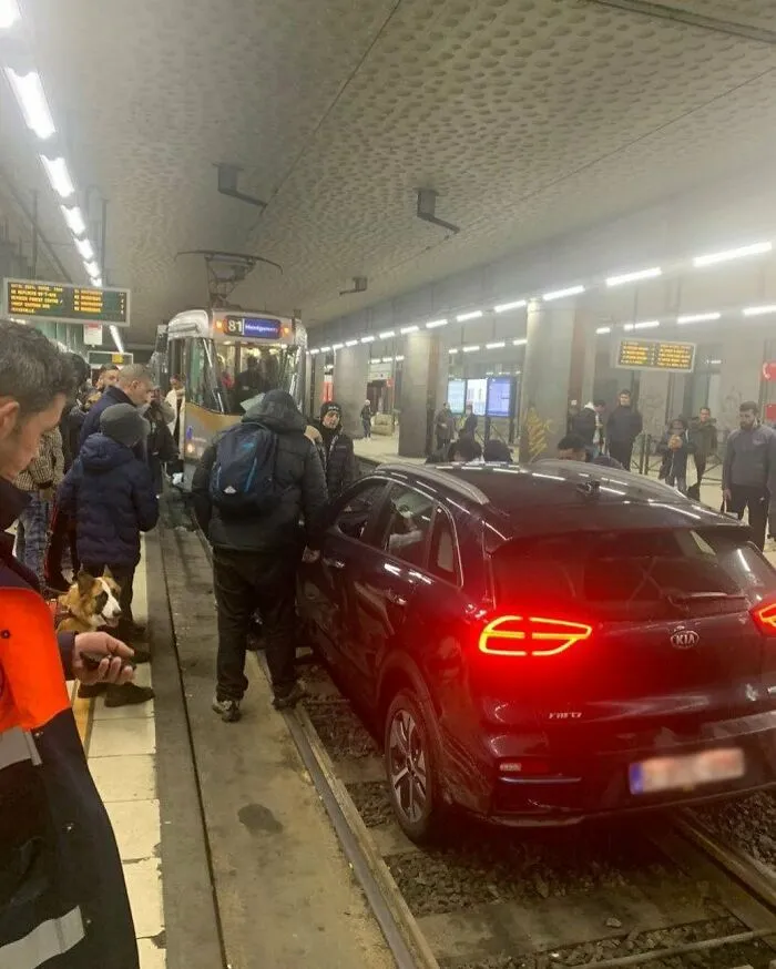 Red car stuck on tram tracks in busy Brussels station with commuters and tram nearby in unusual street scene.