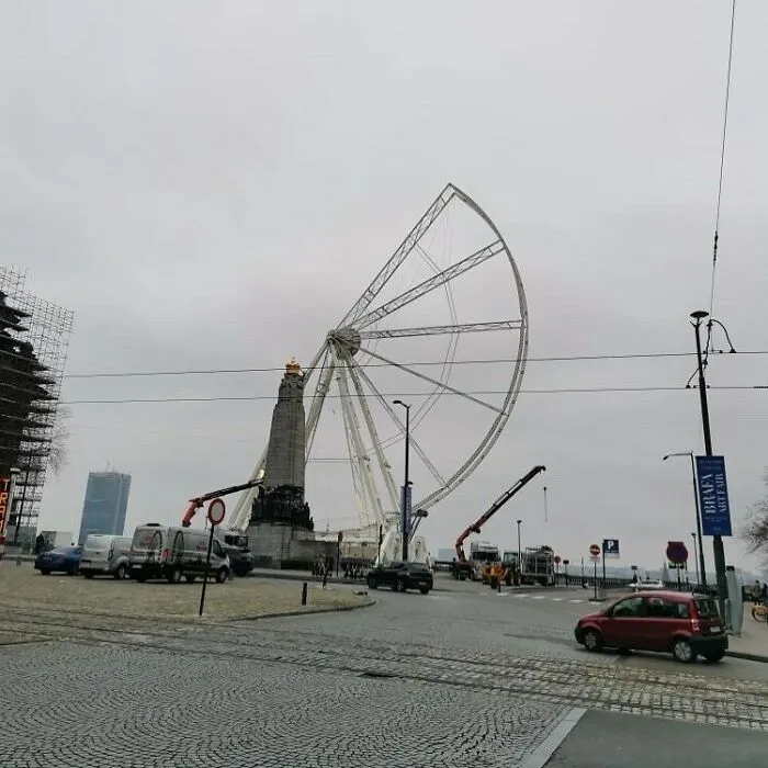 Partially disassembled Ferris wheel near a monument on the streets of Brussels amid urban construction and traffic.