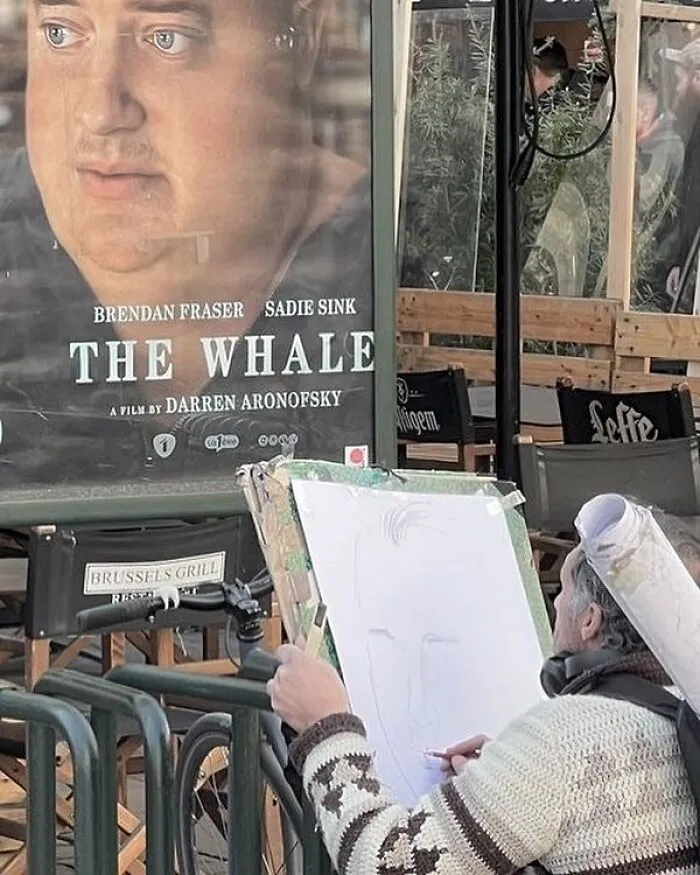 Street artist sketching a portrait near a movie poster in Brussels, showcasing weird and unhinged scenes on the streets.