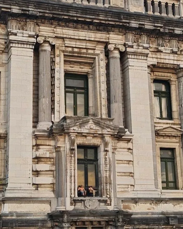 Two people sitting on a narrow ledge of an old ornate building, one of the weirdest things seen on the streets of Brussels.