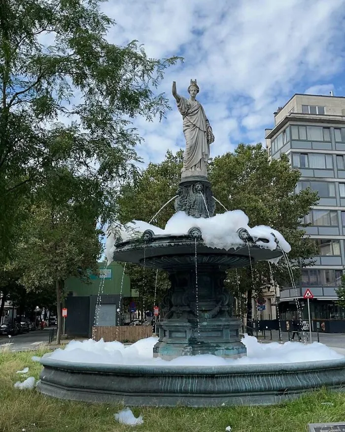 Foamy fountain statue in Brussels street surrounded by trees and buildings, showcasing weird and unhinged city scenes.