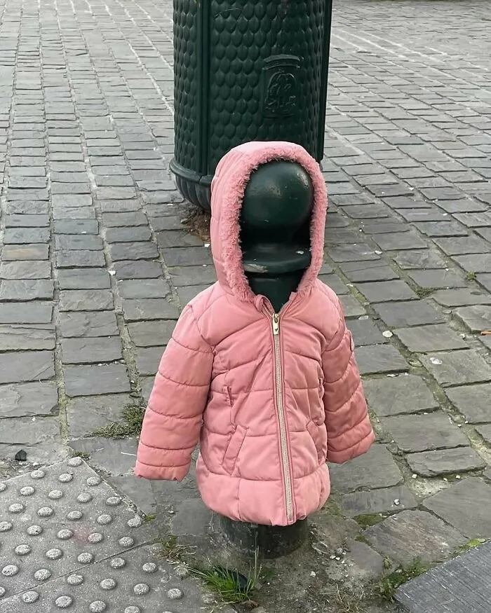 Black street bollard dressed in a pink hooded jacket on cobblestone pavement in Brussels street scene.