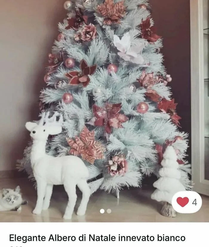 Cat lying near a white Christmas tree decorated with pink flowers and a white reindeer figure indoors.