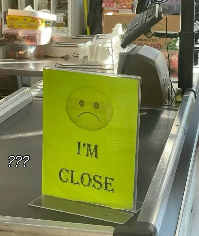 Yellow sign with a sad face emoji and the words "I'M CLOSE" on a store counter, a bizarre meme confusing shoppers.