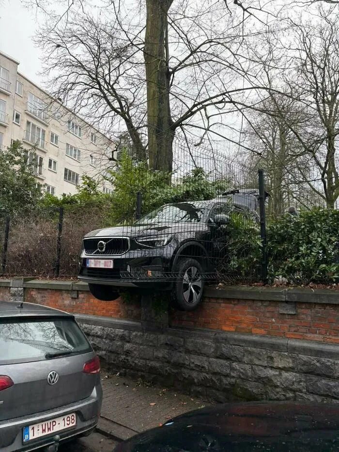 Black Volvo SUV oddly suspended above a wall and caught in a fence, one of the weirdest and most unhinged things in Brussels streets.