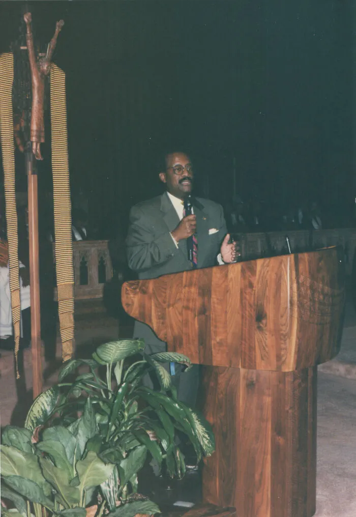 Man in a suit speaking at a wooden podium, sharing local inside jokes with an audience in a dimly lit room.
