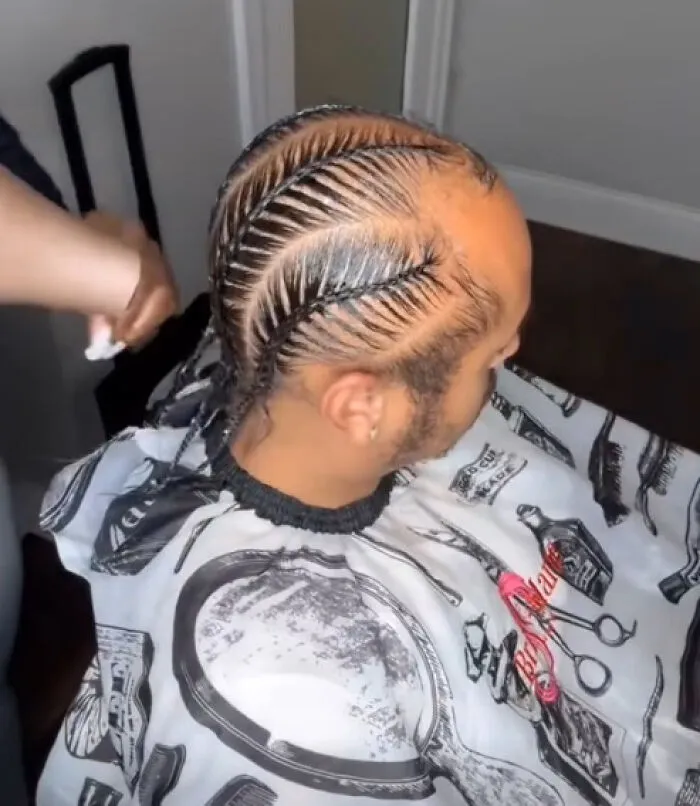 Man with a unique and intricate braided hairstyle sitting in a barber chair wearing a printed haircut cape.
